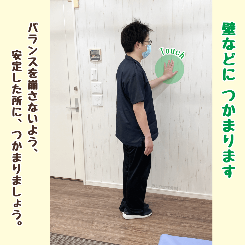 （ステップ1）壁に手をついてバランスをとりながら、立った姿勢で大腿四頭筋ストレッチの準備をしている様子