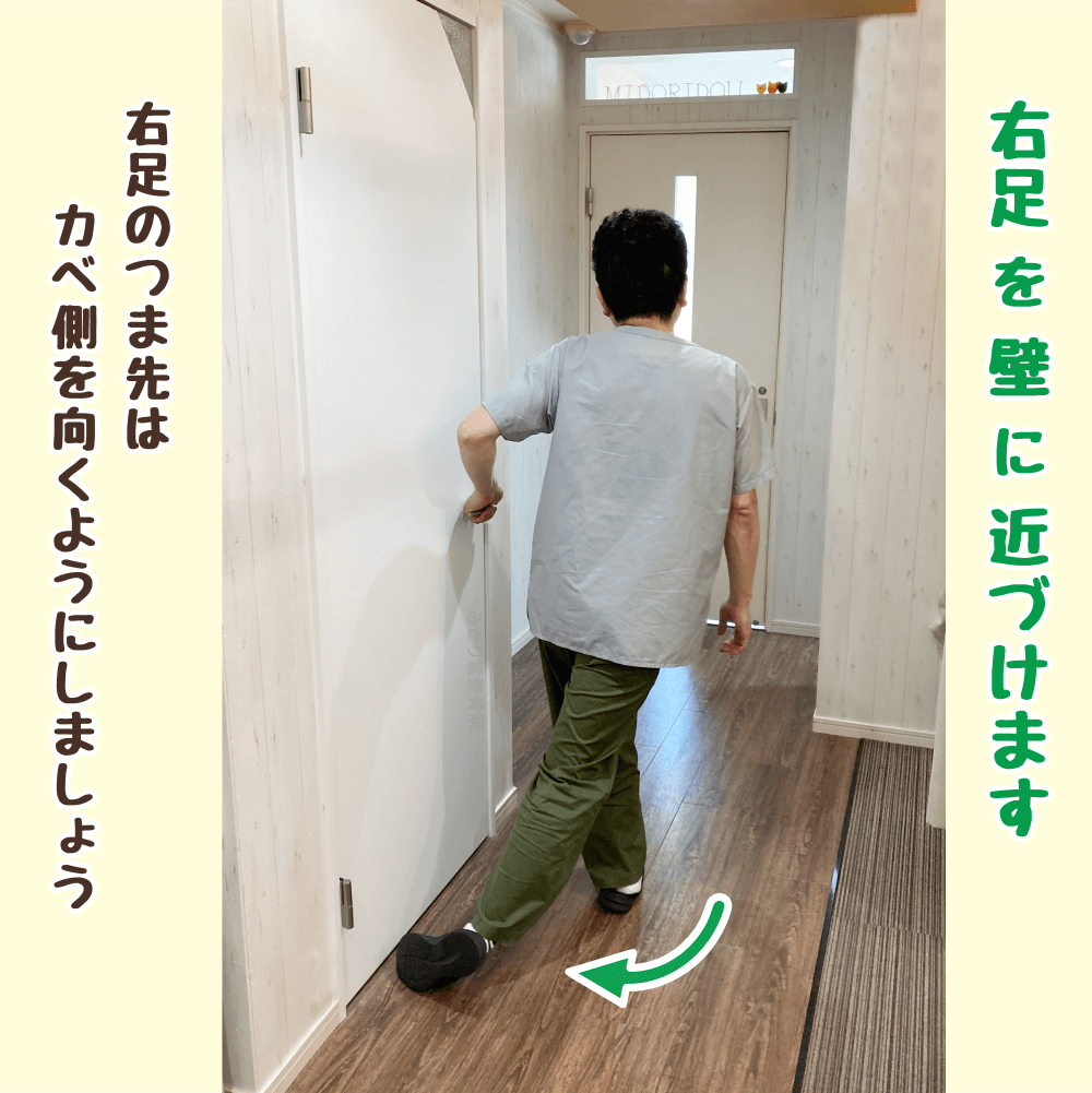右足を後ろに引き、つま先を壁に近づけて姿勢を整える準備運動の途中ステップ。体幹の可動域を広げる動作。